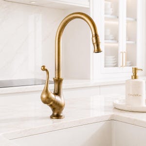 May include: A brass kitchen tap with a high arched spout and a decorative handle. The tap is set against a white countertop and sink. A matching soap dispenser is on the right.