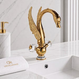 May include: A gold-colored swan-shaped faucet with crystal knobs. The faucet has detailed wing accents and a swan head spout. A white marble soap dispenser and a white towel with the text "ORIVELLEHAUS" are also visible.