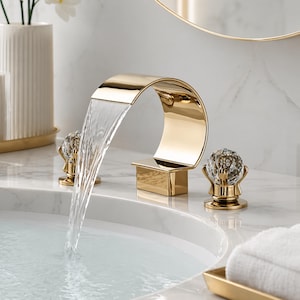 May include: A gold-finished bathroom tap with a waterfall spout. The tap features a curved design and is accompanied by two crystal knobs on gold bases. Water cascades from the spout into a white sink, creating a modern, luxurious look.