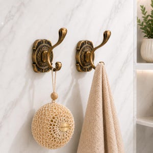 May include: Two antique brass wall hooks with ornate rectangular backplates and curved double prongs. One hook holds a beige mesh bath sponge with a small oval tag reading OrivelleHaus, while the other hook supports a folded beige cotton towel.