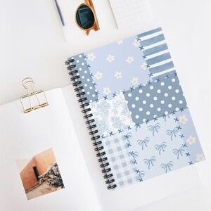May include: A spiral-bound notebook with a light blue patchwork design. The cover features various patterns, including polka dots, stripes, floral prints, and bows. White daisies and blue cross-stitch details add to the design. The notebook is open on a white surface.