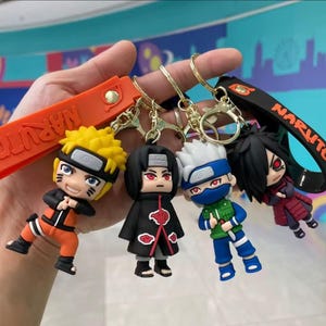 May include: Four character keychains featuring anime figures. The keychains have gold-colored metal rings and clasps. The characters are in various outfits, with orange, black, blue, and red color schemes. One keychain has an orange strap with the word "NARUTO" printed on it.