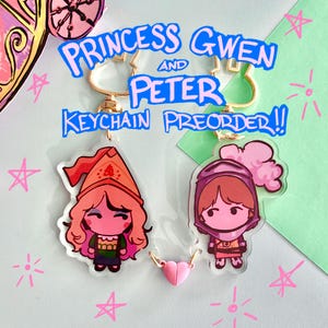 May include: Two acrylic keychains featuring cartoon characters, one in a red hat and the other in a helmet. The keychains are connected by a pink heart charm. The text "PRINCESS GWEN AND PETER KEYCHAIN PREORDER!!" is above the keychains.