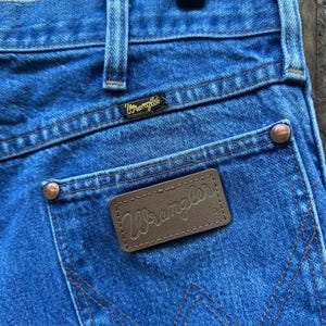 Op de afbeelding: Close-up van een blauwe spijkerbroek met het Wrangler-logo op de achterzak en tailleband. De jeans heeft koperkleurige klinknagels en een bruin leren label met de merknaam Wrangler.