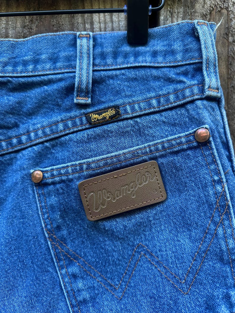 Op de afbeelding: Close-up van een blauwe spijkerbroek met het Wrangler-logo op de achterzak en tailleband. De jeans heeft koperkleurige klinknagels en een bruin leren label met de merknaam Wrangler.
