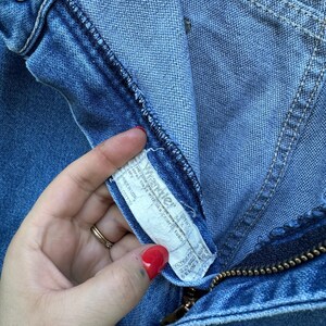 Op de afbeelding: Close-up van een blauwe denim jeans met een messing ritssluiting en een knoopsluiting. Het Wrangler-merklabel is zichtbaar, samen met stikseldetails. De jeans lijken vintage te zijn.