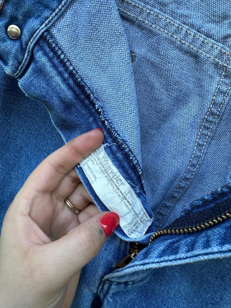 Op de afbeelding: Close-up van een blauwe denim jeans met een messing ritssluiting en een knoopsluiting. Het Wrangler-merklabel is zichtbaar, samen met stikseldetails. De jeans lijken vintage te zijn.
