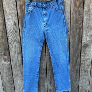Op de afbeelding: Blauwe spijkerbroek hangend aan een houten hanger tegen een verweerde houten achtergrond. De jeans heeft een klassiek ontwerp met vijf zakken, een knoop- en ritssluiting en een rechte pijp. De denim heeft een medium wassing.