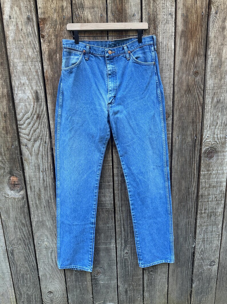 Op de afbeelding: Blauwe spijkerbroek hangend aan een houten hanger tegen een verweerde houten achtergrond. De jeans heeft een klassiek ontwerp met vijf zakken, een knoop- en ritssluiting en een rechte pijp. De denim heeft een medium wassing.