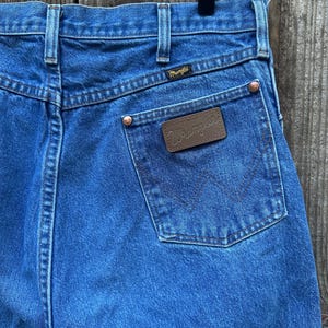 Op de afbeelding: Close-up van een blauwe Wrangler-jeans van denim. De jeans heeft een bruine leren patch met het Wrangler-logo op de achterzak. De achterzakken hebben een kenmerkende W-vormige stiksels. De jeans heeft koperen klinknagels en een zwart label.