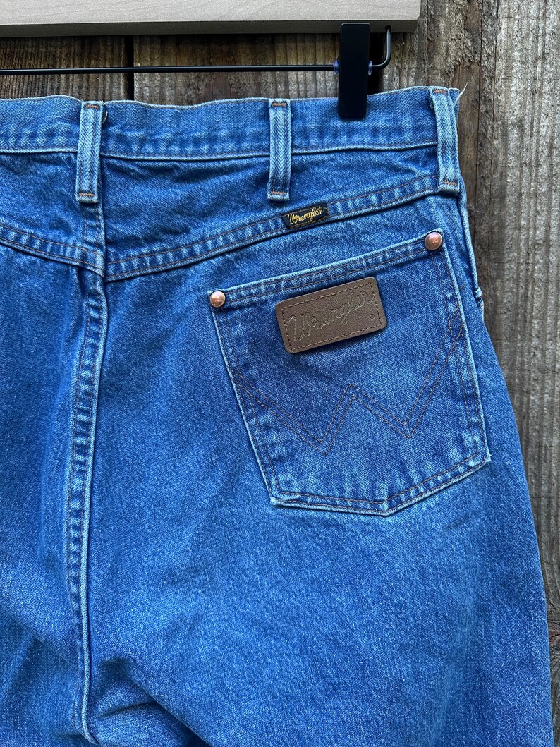 Op de afbeelding: Close-up van een blauwe Wrangler-jeans van denim. De jeans heeft een bruine leren patch met het Wrangler-logo op de achterzak. De achterzakken hebben een kenmerkende W-vormige stiksels. De jeans heeft koperen klinknagels en een zwart label.