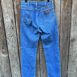 Op de afbeelding: Blauwe denim jeans hangen tegen een houten achtergrond. De jeans hebben een klassiek ontwerp met vijf zakken, met een bruine leren patch op de achterzak. Het Wrangler-logo is zichtbaar. De jeans hebben een medium wassing en lijken vintage.