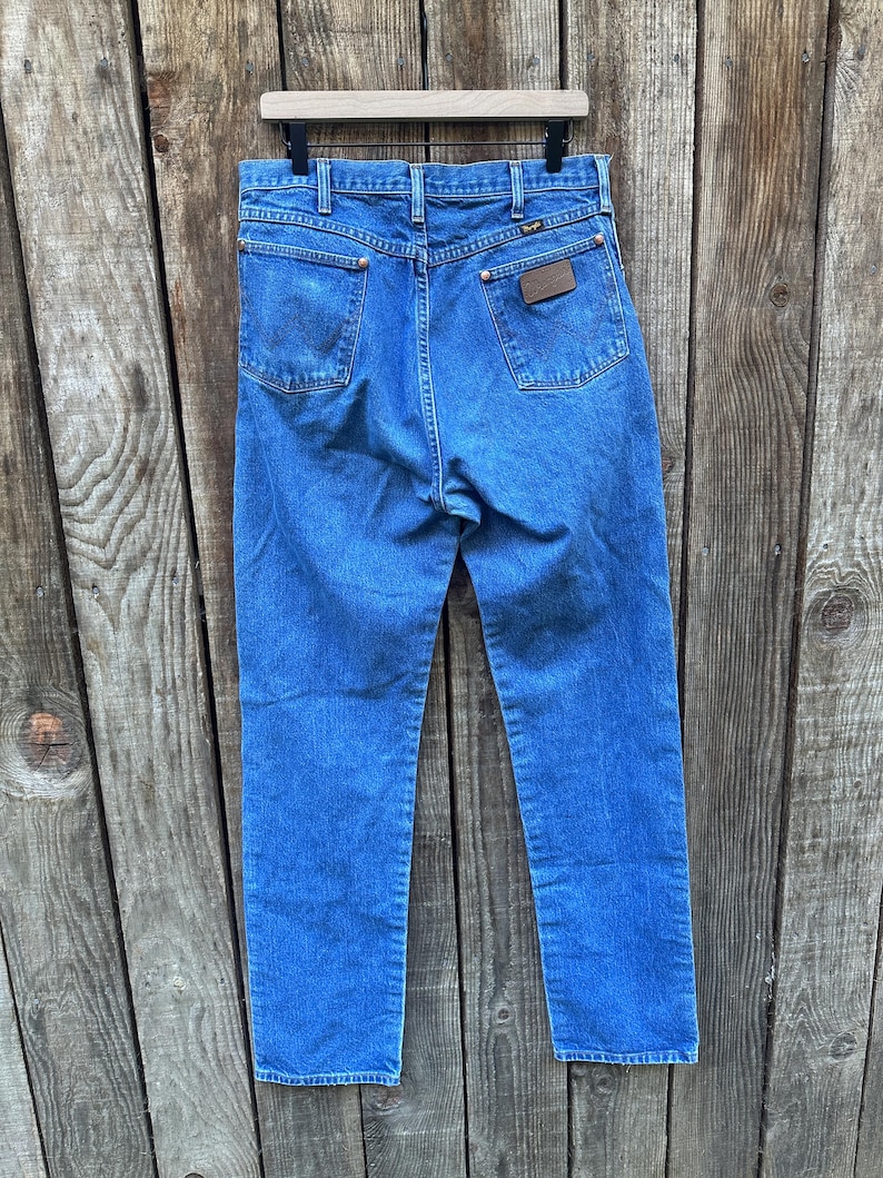 Op de afbeelding: Blauwe denim jeans hangen tegen een houten achtergrond. De jeans hebben een klassiek ontwerp met vijf zakken, met een bruine leren patch op de achterzak. Het Wrangler-logo is zichtbaar. De jeans hebben een medium wassing en lijken vintage.
