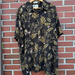 May include: A black short-sleeve button-down shirt featuring a repeating pattern of martini glasses and tropical palm leaves in shades of yellow, green, and white. The shirt has a standard collar, a front button placket, and a single chest pocket.
