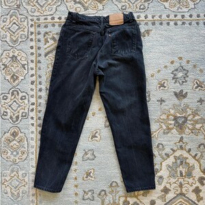 Peut inclure: Un jean en denim noir avec une taille haute, une étiquette en cuir beige sur la ceinture arrière, deux poches arrière, des passants de ceinture et une petite étiquette orange sur la poche arrière droite, vu de dos.
