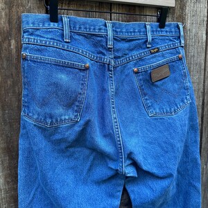 Op de afbeelding: Blauwe denim jeans met het Wrangler-logo op de achterzak. De jeans hangen aan een houten kleerhanger. De jeans hebben twee achterzakken en koperkleurige klinknagels. De denim heeft een medium wassing.