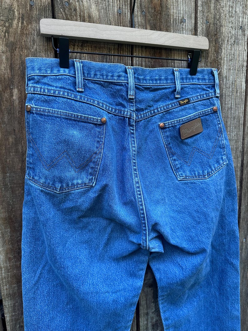 Op de afbeelding: Blauwe denim jeans met het Wrangler-logo op de achterzak. De jeans hangen aan een houten kleerhanger. De jeans hebben twee achterzakken en koperkleurige klinknagels. De denim heeft een medium wassing.