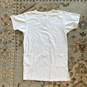 Op de afbeelding: Een wit T-shirt-jurkje met korte mouwen en een decoratief uitgesneden kanten patroon rond de halslijn. Het kledingstuk heeft twee vierkante opgestikte zakken aan de voorzijde, elk versierd met kleine metalen studs, en zijsplitten bij de zoom.