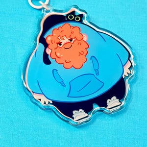 Könnte beinhalten: Ein klarer Acryl-Anhänger mit einer Cartoonfigur in einem blauen Kapuzenpullover mit orangefarbenem Haar und einem roten Bart. Der Anhänger hat einen weißen Verschluss und einen weißen Ring. Die Figur trägt dunkelblaue Hosen und froschförmige Schuhe.