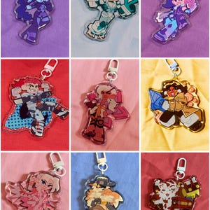 Puede incluir: Una colecci&oacute;n de nueve llaveros de acr&iacute;lico con varios personajes ilustrados en colores vibrantes, cada uno unido a un cierre de pl&aacute;stico. Los dise&ntilde;os incluyen figuras con armaduras, atuendos estilizados y poses &uacute;nicas.