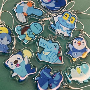 Puede incluir: Una colección de colgantes de acrílico transparente con varias criaturas de dibujos animados en tonos azules, blancos y amarillos. Cada colgante tiene una cuerda blanca para colgar. Los diseños incluyen una tortuga, una rana, un pingüino y otros personajes acuáticos.