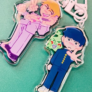 Pode incluir: Dois chaveiros de acrílico transparente com personagens de anime. Um chaveiro mostra um personagem loiro em um terno roxo claro, fazendo o sinal de paz. O outro mostra um personagem em uniforme azul com uma criatura verde. Ambos os chaveiros têm ferragens brancas.