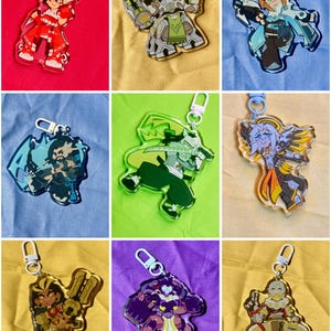 Puede incluir: Una colecci&oacute;n de nueve llaveros de acr&iacute;lico con diversos personajes ilustrados en varias poses y esquemas de color, cada uno unido a un cierre giratorio de pl&aacute;stico blanco o azul claro, mostrando dise&ntilde;os gr&aacute;ficos detallados y definidos.