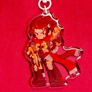 Puede incluir: Un llavero de acr&iacute;lico con un personaje de dibujos animados estilizado de pelo rojo puntiagudo, con armadura roja y amarilla y un arma futurista amarilla. El personaje lleva una larga bufanda roja y tiene un cierre giratorio de pl&aacute;stico blanco.