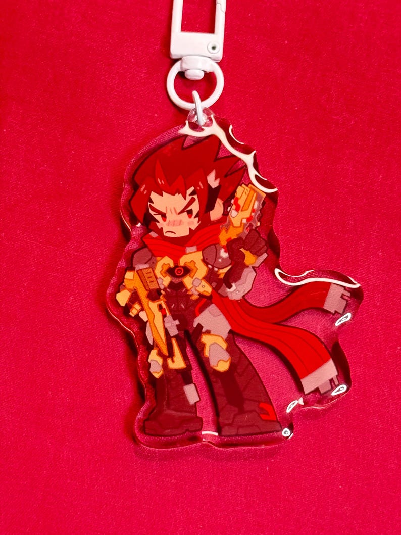 Puede incluir: Un llavero de acr&iacute;lico con un personaje de dibujos animados estilizado de pelo rojo puntiagudo, con armadura roja y amarilla y un arma futurista amarilla. El personaje lleva una larga bufanda roja y tiene un cierre giratorio de pl&aacute;stico blanco.
