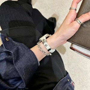 May include: A white watch strap featuring metallic star-shaped studs and a silver-toned chain link bracelet. The watch strap is secured with a buckle and is worn alongside two silver rings on the fingers, showcasing a coordinated metallic aesthetic.