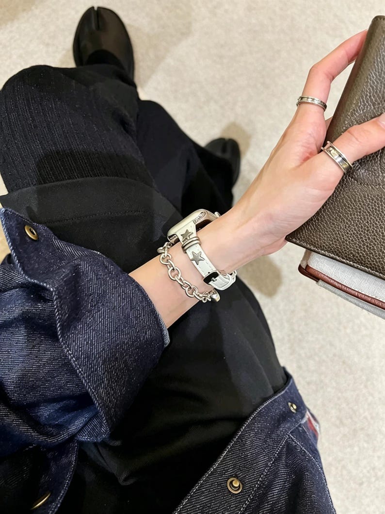 May include: A white watch strap featuring metallic star-shaped studs and a silver-toned chain link bracelet. The watch strap is secured with a buckle and is worn alongside two silver rings on the fingers, showcasing a coordinated metallic aesthetic.