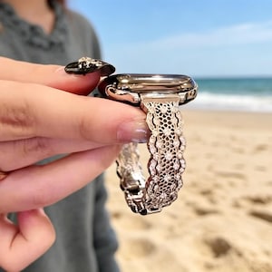 May include: A silver-toned smartwatch with a decorative, openwork band featuring small, sparkling embellishments. The watch is held up against a beach backdrop, with the ocean and sky visible in the background. A gold ring is visible on the person's finger.