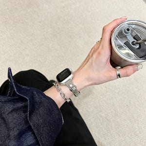 May include: A smartwatch with a dark screen and a grey strap featuring a star pattern, worn alongside a silver chain link bracelet. The watch is paired with a silver ring and a metallic tumbler with a black lid, worn with a dark denim jacket.