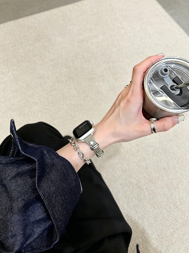 May include: A smartwatch with a dark screen and a grey strap featuring a star pattern, worn alongside a silver chain link bracelet. The watch is paired with a silver ring and a metallic tumbler with a black lid, worn with a dark denim jacket.