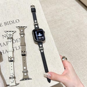 May include: Three slim watch bands for a smartwatch, in white, beige, and black, each featuring silver-toned star-shaped studs and metal loops. The black band is attached to a smartwatch displaying a digital clock face with the time 20:16.