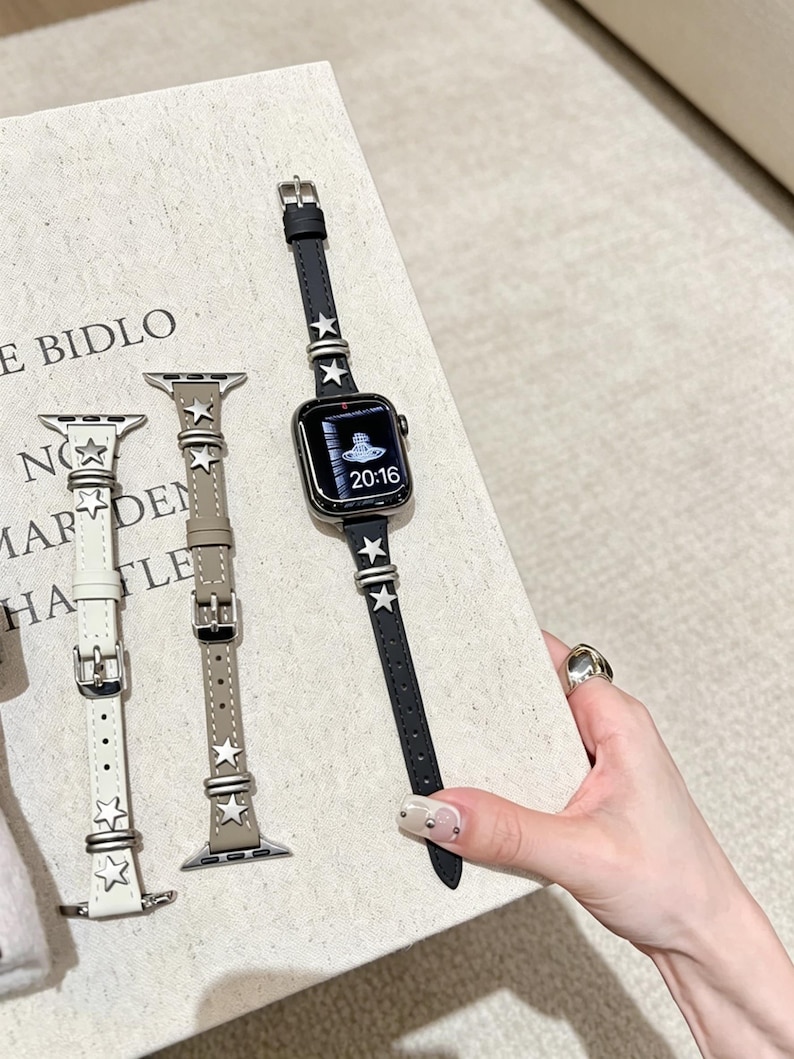 May include: Three slim watch bands for a smartwatch, in white, beige, and black, each featuring silver-toned star-shaped studs and metal loops. The black band is attached to a smartwatch displaying a digital clock face with the time 20:16.