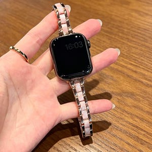 May include: A rose gold smartwatch with a pink and rose gold band. The watch face displays the time 16:03. The band is made of rectangular links. A gold ring is visible on the left hand.