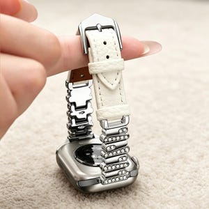 Op de afbeelding: Een zilverkleurige smartwatch-behuizing met een witte, getextureerde leren band. De band is voorzien van een zilveren gesp en een reeks zilverkleurige metalen schakels met kleine heldere kristallen die het leer met de behuizing verbinden.
