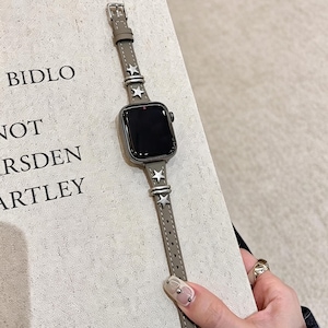 May include: A taupe leather watch band for a smartwatch featuring silver-toned star-shaped metal studs and matching silver-toned metal loops. The band is attached to a rectangular smartwatch with a black screen, showcasing a sleek and functional design.