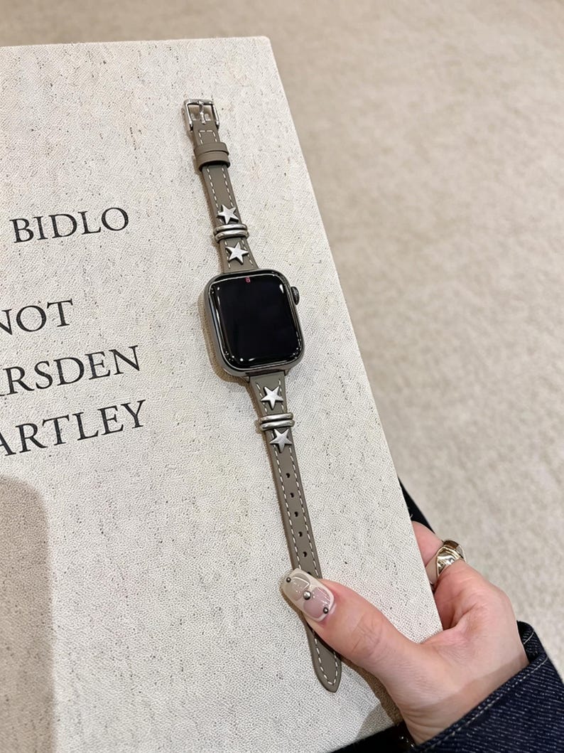 May include: A taupe leather watch band for a smartwatch featuring silver-toned star-shaped metal studs and matching silver-toned metal loops. The band is attached to a rectangular smartwatch with a black screen, showcasing a sleek and functional design.