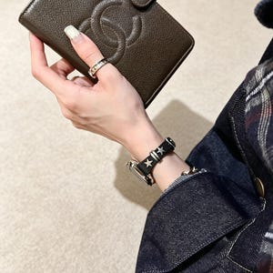 May include: A dark brown textured leather wallet featuring a prominent embossed interlocking logo and a snap closure. The wallet is held alongside a black watch strap adorned with silver-toned star-shaped studs and a metallic ring on the finger.