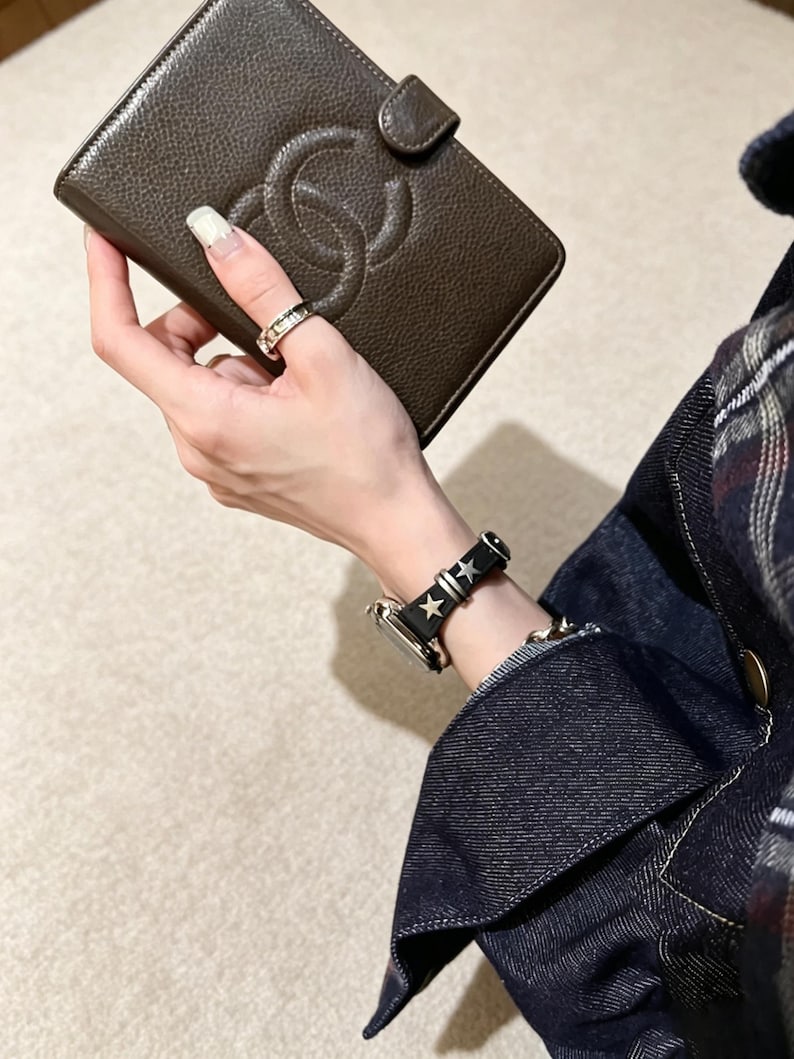 May include: A dark brown textured leather wallet featuring a prominent embossed interlocking logo and a snap closure. The wallet is held alongside a black watch strap adorned with silver-toned star-shaped studs and a metallic ring on the finger.
