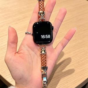May include: A black smartwatch with a brown braided leather band and gold-colored metal accents. The watch face displays the time 16:58. The band features a clasp closure and decorative elements.