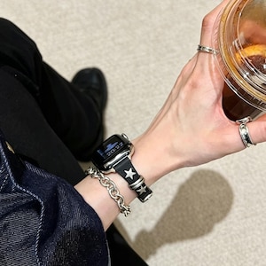 May include: A black smartwatch band featuring silver-toned star-shaped studs and metallic loops. The band is attached to a digital watch face displaying the time 20:22. A thick silver-toned chain bracelet is worn on the same wrist.