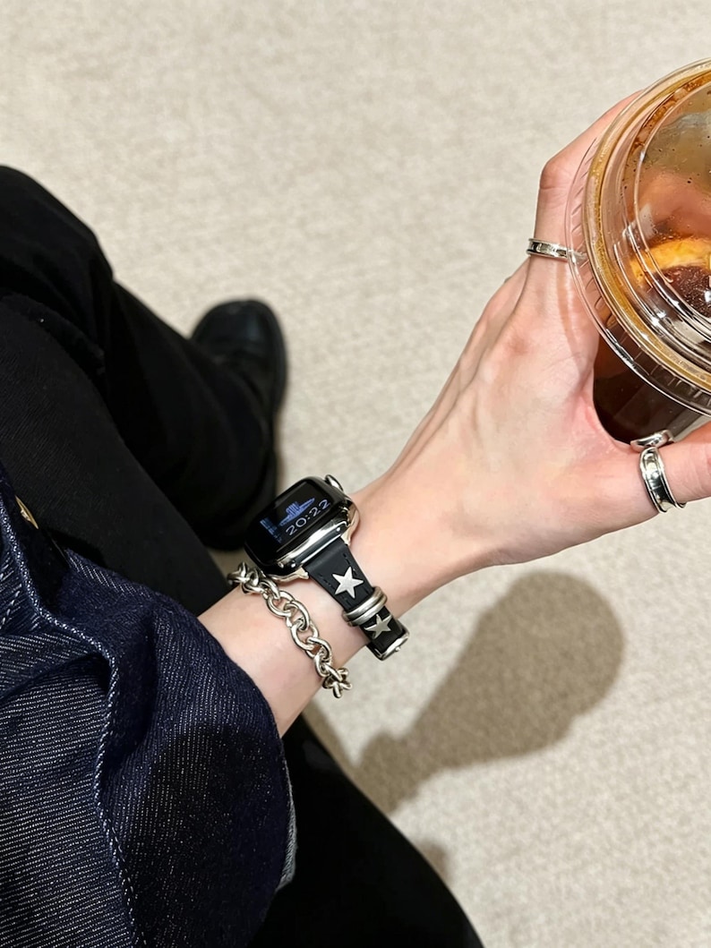 May include: A black smartwatch band featuring silver-toned star-shaped studs and metallic loops. The band is attached to a digital watch face displaying the time 20:22. A thick silver-toned chain bracelet is worn on the same wrist.