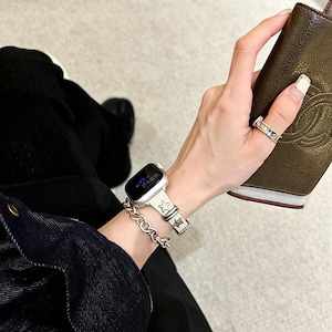 May include: A silver-toned smartwatch band featuring a repeating star pattern, paired with a matching silver-toned chain link bracelet. The watch face is black with digital text displaying the time, and a silver ring is worn on the finger.
