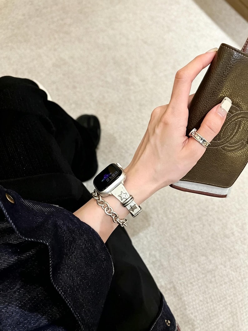 May include: A silver-toned smartwatch band featuring a repeating star pattern, paired with a matching silver-toned chain link bracelet. The watch face is black with digital text displaying the time, and a silver ring is worn on the finger.