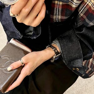 May include: A black leather watch band featuring silver-toned star-shaped studs and a metal buckle, attached to a digital watch. The band is worn on a wrist alongside a brown textured wallet with a logo and a plaid denim jacket with dark blue fabric.