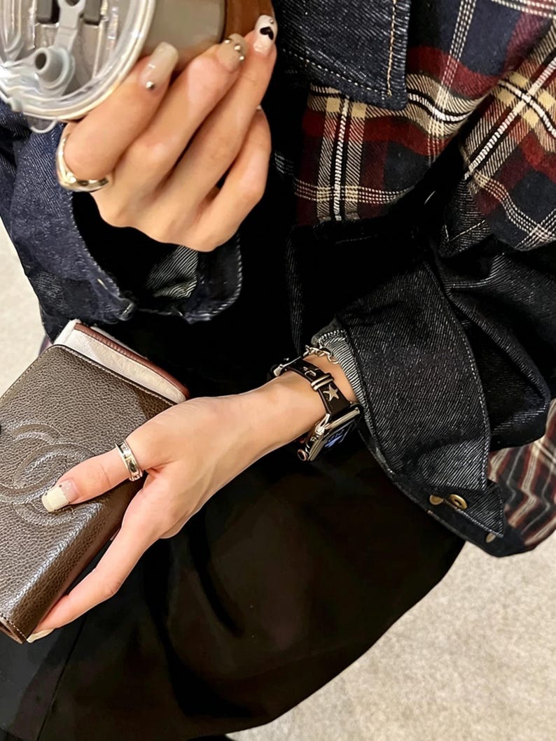May include: A black leather watch band featuring silver-toned star-shaped studs and a metal buckle, attached to a digital watch. The band is worn on a wrist alongside a brown textured wallet with a logo and a plaid denim jacket with dark blue fabric.