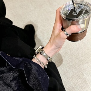 May include: A smartwatch with a grey leather strap featuring silver star studs, paired with a silver-toned chain link bracelet. The watch is worn on a wrist alongside a silver ring, while holding a stainless steel tumbler with a dark wood accent.